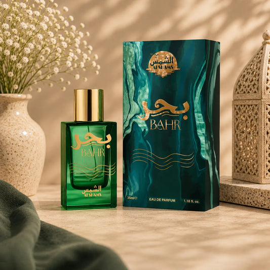 A green glass perfume bottle with a gold cap stands beside its matching teal and gold box on a warm beige surface. Decorative flowers in a vase, draped fabric, and a lantern are visible around it.