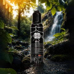 A black cylindrical bottle with a black cap stands centered in a forest stream scene with a waterfall and green leaves in the background. The bottle has a glossy black finish with a white spiderweb graphic and the brand name 'A MIRA' visible on the front.