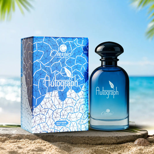 A blue perfume bottle labeled 'Autograph Classic' stands beside its matching blue and white patterned box on a stone ledge at the beach, with ocean and palm leaves in the background.