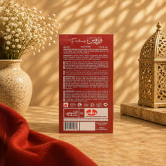 The back of a red Firdaus by Al Mahara perfume box, displayed on a wooden surface with a red fabric cloth, a ceramic vase of dried flowers, and a decorative brass lantern in the background.