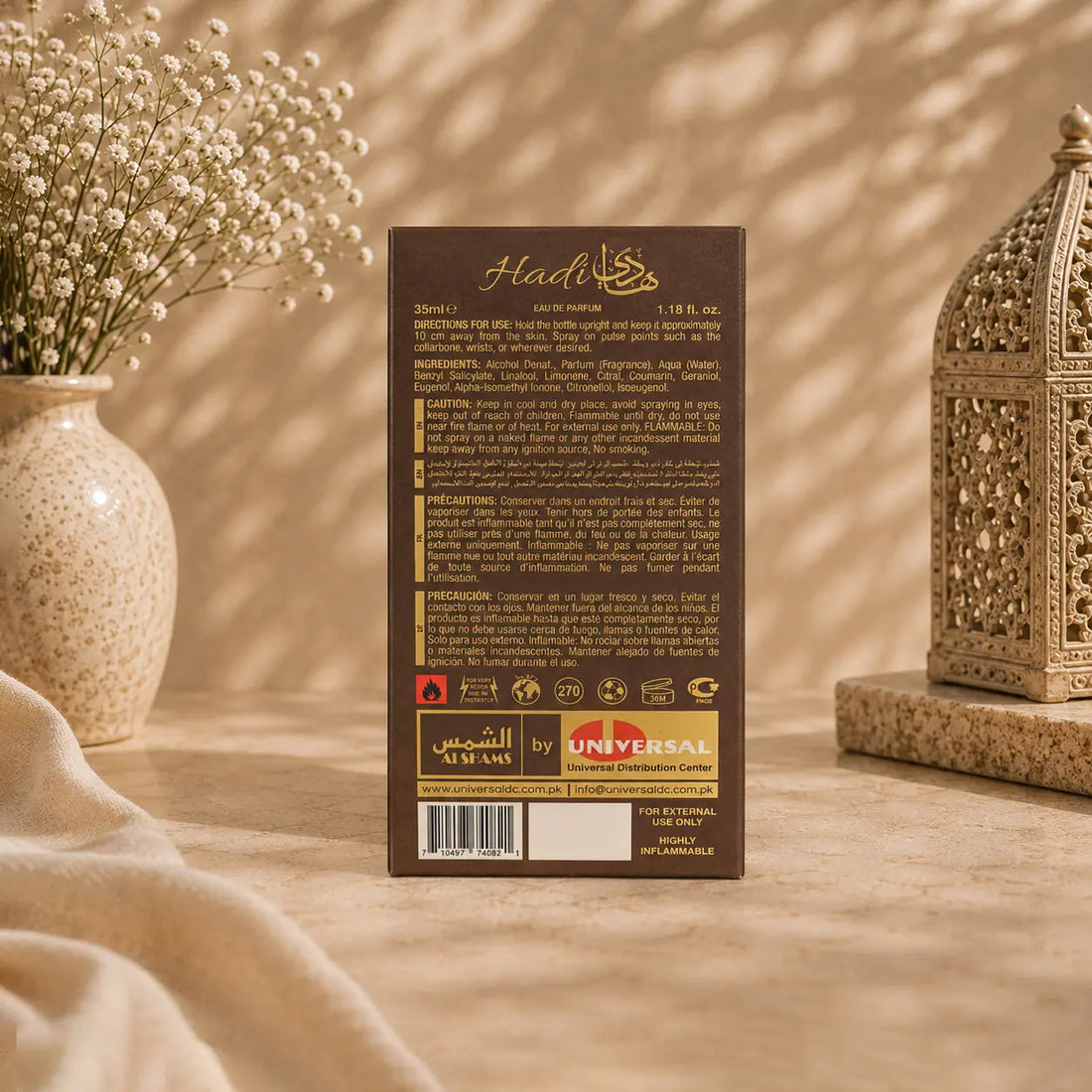A 30cl pack of Hadh chocolate spread, showing the back of the brown cardboard packaging with gold text and logos, styled on a beige surface with a vase of baby's breath and a decorative lantern in the background.