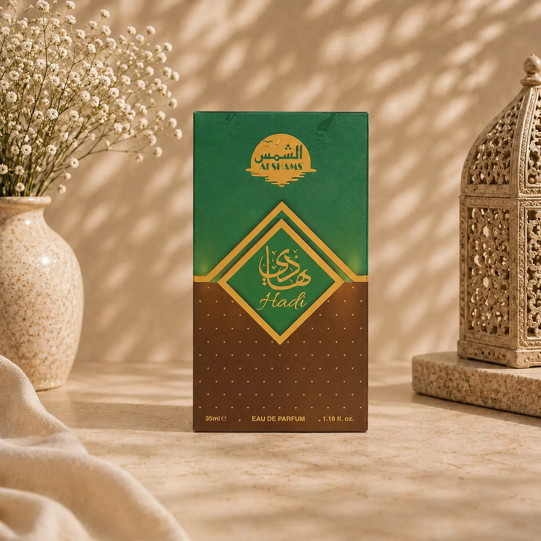 A rectangular perfume box standing upright on a beige surface, with a green-to-brown gradient front panel and gold Arabic lettering. The box is styled in a warm indoor setting with a vase of small white flowers on the left and an ornate lantern on the right.