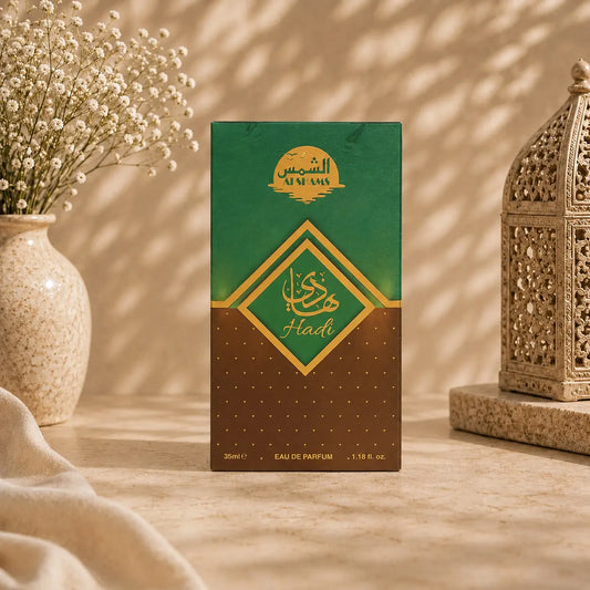 A rectangular perfume box standing upright on a beige surface, with a green-to-brown gradient front panel and gold Arabic lettering. The box is styled in a warm indoor setting with a vase of small white flowers on the left and an ornate lantern on the right.