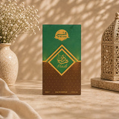 A rectangular perfume box standing upright on a beige surface, with a green-to-brown gradient front panel and gold Arabic lettering. The box is styled in a warm indoor setting with a vase of small white flowers on the left and an ornate lantern on the right.