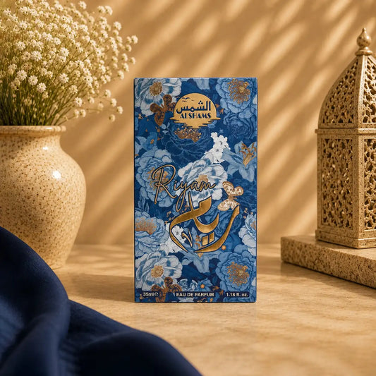 A rectangular perfume box standing upright on a tabletop, decorated with a blue floral pattern and gold Arabic script. The box is photographed in a styled interior scene with a vase of small white flowers on the left, a gold lantern on the right, and a dark navy fabric cloth in the foreground.