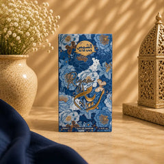 A rectangular perfume box standing upright on a tabletop, decorated with a blue floral pattern and gold Arabic script. The box is photographed in a styled interior scene with a vase of small white flowers on the left, a gold lantern on the right, and a dark navy fabric cloth in the foreground.