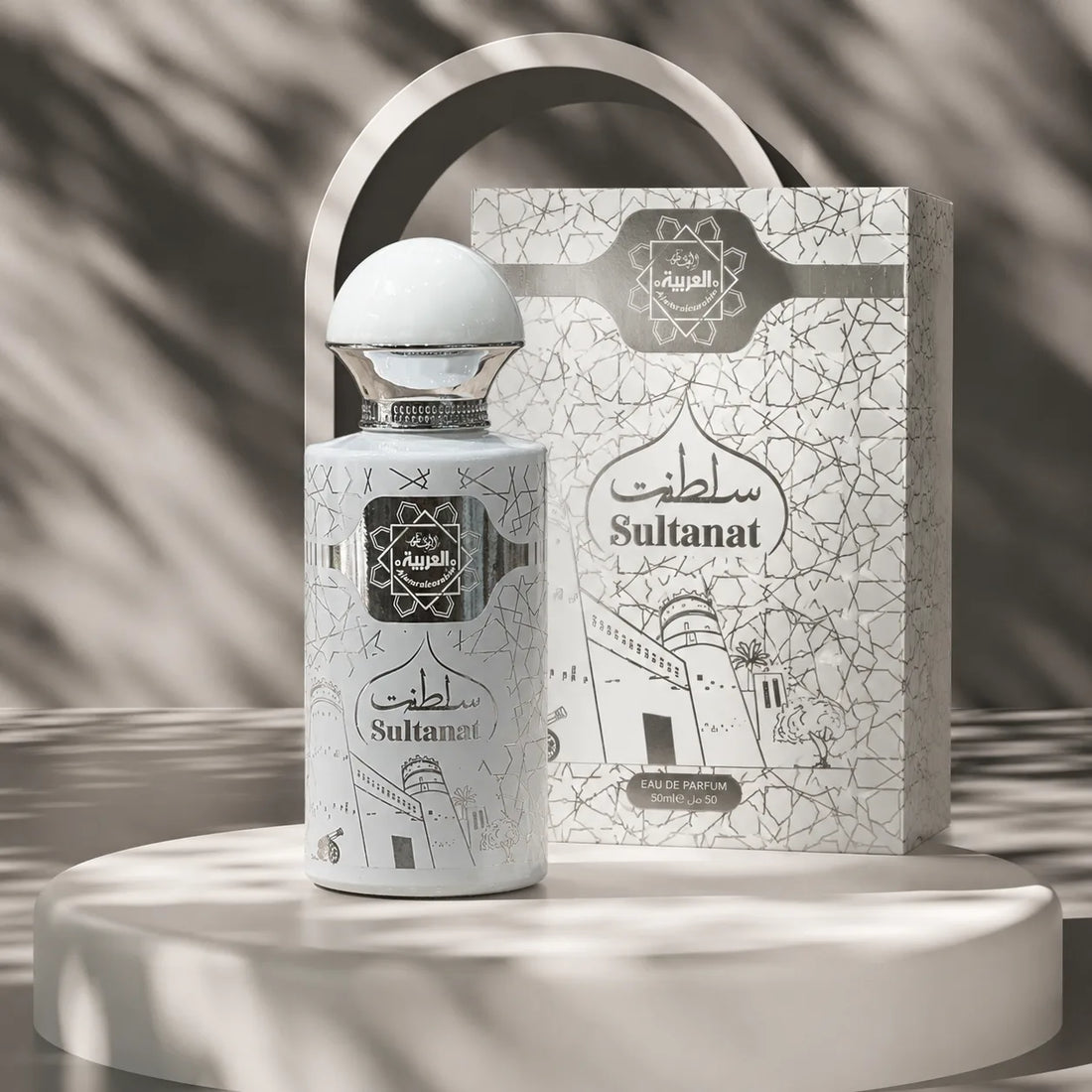 A white perfume bottle with a silver cap stands in front of its matching illustrated box on a white pedestal. The bottle and box are decorated with black architectural line drawings and the words 'Sultanat' and 'Al-Ahlia'.