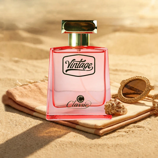 A pink rectangular perfume bottle with a gold cap sits on a folded beige towel on a sandy beach. A small seashell and a gold-framed mirror are placed beside it.